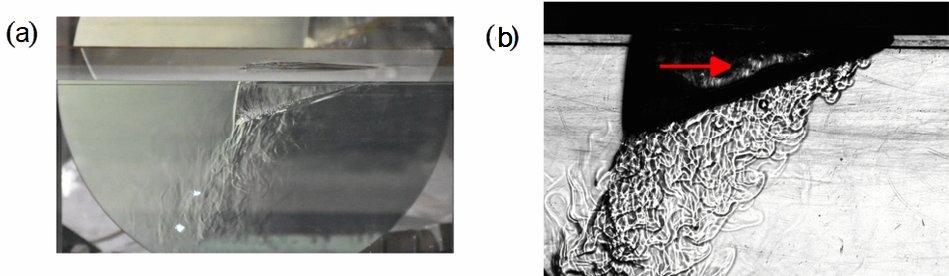 Fig. 3. Vue du montage d’ombroscopie et l’image obtenue a) Vue de profil du bloc de glace flottant avec un miroir parabolique installé derrière le récipient d’eau douce. L’eau froide plus dense engendre un flux de convection qui est visible sous le bloc de glace. Le miroir est utilisé pour former l’image en ombroscopie b) Image en ombroscopie qui montre le glaçon se déplaçant vers la droite avec le flux de convection vers l’arrière du bloc. M. Berhanu et al. Physical Review Fluids 11, March 2026, avec autorisation.